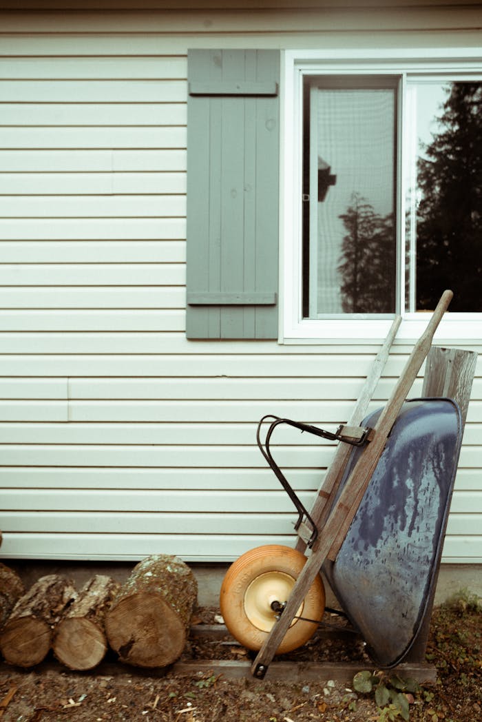 A rustic wheelbarrow leaning against a house with logs, featuring modern siding and a window.